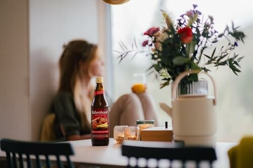 Frau schaut aus dem Fenster neben Blumenstrauß und Saftflasche.